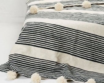 May include: A white and black striped throw blanket with white pom-poms along the edges. The blanket is folded on a bed with two white pillows with pom-poms.