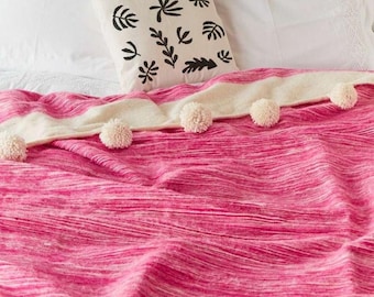 May include: A pink and white striped throw blanket with white pom-poms along the edge. The blanket is folded on a bed with white pillows and a white pillow with a black leaf pattern.