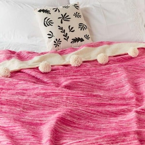 Könnte beinhalten: Eine gestreifte rosa und weiße Wolldecke mit weißen Bommeln am Rand. Die Decke liegt auf einem Bett mit weißen Kissen und einem weißen Kissen mit einem schwarzen Blattmotiv.