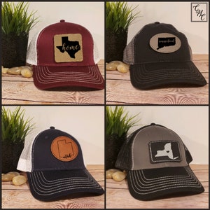 May include: Four trucker hats with different state outlines and names on leather patches. The hats are all black with white mesh back panels. The patches are brown leather with black state outlines and white text. The states are Texas, Connecticut, Utah, and New York.