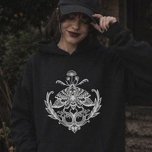 K&ouml;nnte beinhalten: Ein schwarzer Kapuzenpullover mit einem wei&szlig;en Grafikdruck auf der Brust. Das Design zeigt einen Nachtfalter &uuml;ber zwei Schlangen, die mit belaubten Zweigen verschlungen sind, gekr&ouml;nt von einem kleinen Pilz. Der Hoodie hat lange &Auml;rmel und eine Tasche.