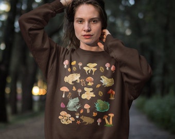 May include: A dark brown long-sleeve sweatshirt featuring a graphic print of various colorful mushrooms and frogs. The design includes multiple species of fungi in shades of orange, red, and tan, alongside several frogs in green and brown tones.