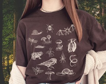 May include: A dark brown t-shirt featuring a detailed white illustration of various forest creatures and plants, including a snail, bat, mushrooms, and a snake. The shirt is worn with blue jeans and a cream-colored cardigan.