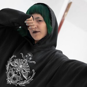K&ouml;nnte beinhalten: Ein schwarzer Kapuzenpullover mit einem wei&szlig;en Grafikdruck auf der Brust. Das Design zeigt einen detaillierten Nachtfalter, umgeben von botanischen Elementen wie kleinen Pilzen und bl&auml;ttrigen Ranken, dargestellt in einem klaren Linienstil.