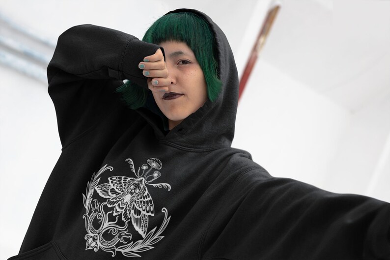 K&ouml;nnte beinhalten: Ein schwarzer Kapuzenpullover mit einem wei&szlig;en Grafikdruck auf der Brust. Das Design zeigt einen detaillierten Nachtfalter, umgeben von botanischen Elementen wie kleinen Pilzen und bl&auml;ttrigen Ranken, dargestellt in einem klaren Linienstil.