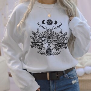 May include: White sweatshirt with a black and white graphic print of a moth, a pentagram, moons, and a bottle. The graphic is centered on the shirt.