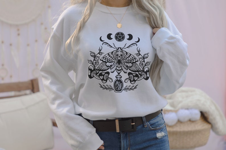May include: White sweatshirt with a black and white graphic print of a moth, a pentagram, moons, and a bottle. The graphic is centered on the shirt.