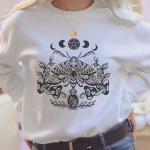 May include: White sweatshirt with a black and white graphic print of a moth, mushrooms, and a bottle. The graphic also includes a pentagram and three crescent moons.