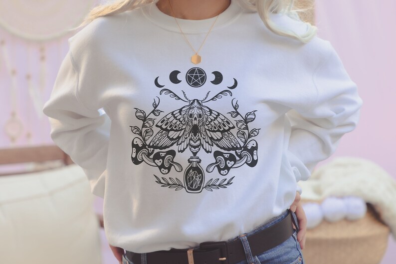 May include: White sweatshirt with a black and white graphic print of a moth, mushrooms, and a bottle. The graphic also includes a pentagram and three crescent moons.