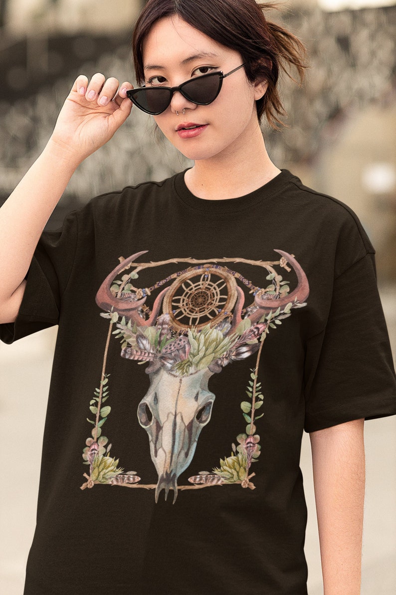 May include: Black t-shirt featuring a detailed illustration of a skull with antlers, a dreamcatcher, and floral accents. The design incorporates earthy tones of brown, green, and cream, creating a bohemian aesthetic. The shirt is a classic crew neck.
