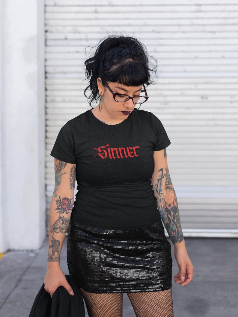May include: A black t-shirt with the word "Sinner" in red gothic lettering. The person wearing the shirt has black hair, black framed glasses, and tattoos on their arms.