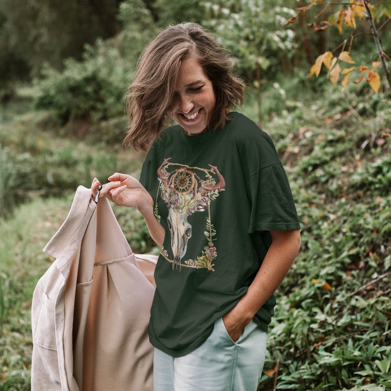 May include: Dark green t-shirt featuring a detailed graphic of a skull with horns, a dreamcatcher, and floral accents. The design is framed with a gold-colored border. The shirt is worn by a person in an outdoor setting.