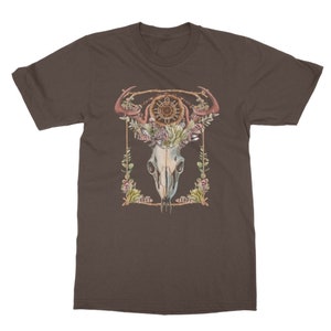 May include: Brown t-shirt featuring a watercolor design of a skull with antlers, a dreamcatcher, and floral elements. The design is framed with a gold border and green leaves. The shirt is made of a soft material.