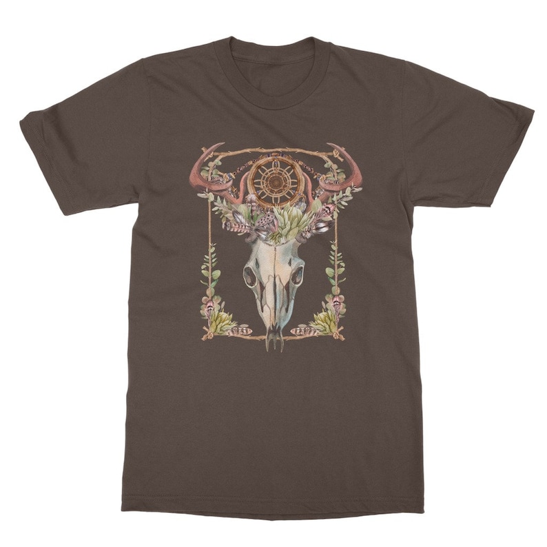 May include: Brown t-shirt featuring a watercolor design of a skull with antlers, a dreamcatcher, and floral elements. The design is framed with a gold border and green leaves. The shirt is made of a soft material.