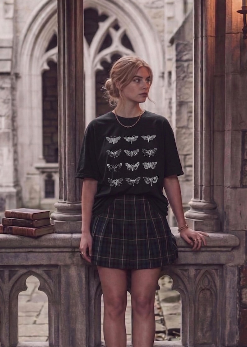 Op de afbeelding: Een zwart t-shirt met een wit motontwerp en een groene geruite rok. Het t-shirt heeft een raster van motillustraties. De rok heeft een groen, zwart en wit ruitpatroon. De outfit wordt gedragen in een stenen gebouw.