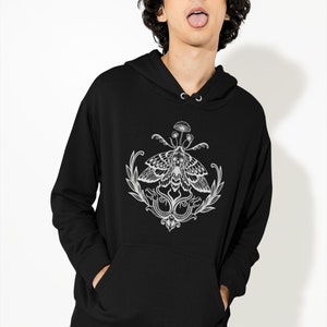 K&ouml;nnte beinhalten: Ein schwarzer Kapuzenpullover mit einem wei&szlig;en Grafikdruck auf der Brust. Das Design zeigt einen Nachtfalter mit einem Totenkopfmuster auf den Fl&uuml;geln, gekr&ouml;nt von einem kleinen Pilz und flankiert von zwei Schlangen und Blattzweigen.