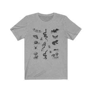 May include: A heather gray t-shirt featuring a detailed black and white illustration of various insects, amphibians, and fungi. The design includes bats, snakes, moths, a frog, a snail, and mushrooms. The shirt has a crew neck and short sleeves.