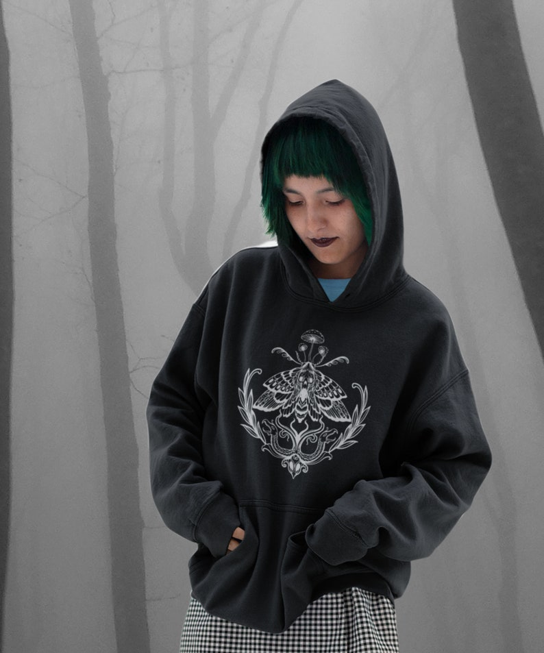 K&ouml;nnte beinhalten: Ein anthrazitfarbener Kapuzenpullover mit einem wei&szlig;en Grafikdruck auf der Brust. Das Design zeigt einen detaillierten Nachtfalter &uuml;ber einem symmetrischen botanischen Arrangement mit kleinen Pilzen, umrahmt von geschwungenen Bl&auml;ttern.