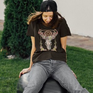 May include: A black t-shirt featuring a detailed graphic of a steer skull adorned with floral elements and a dreamcatcher design. The shirt is worn with gray jeans and a black baseball cap. The image is set outdoors with a green background.