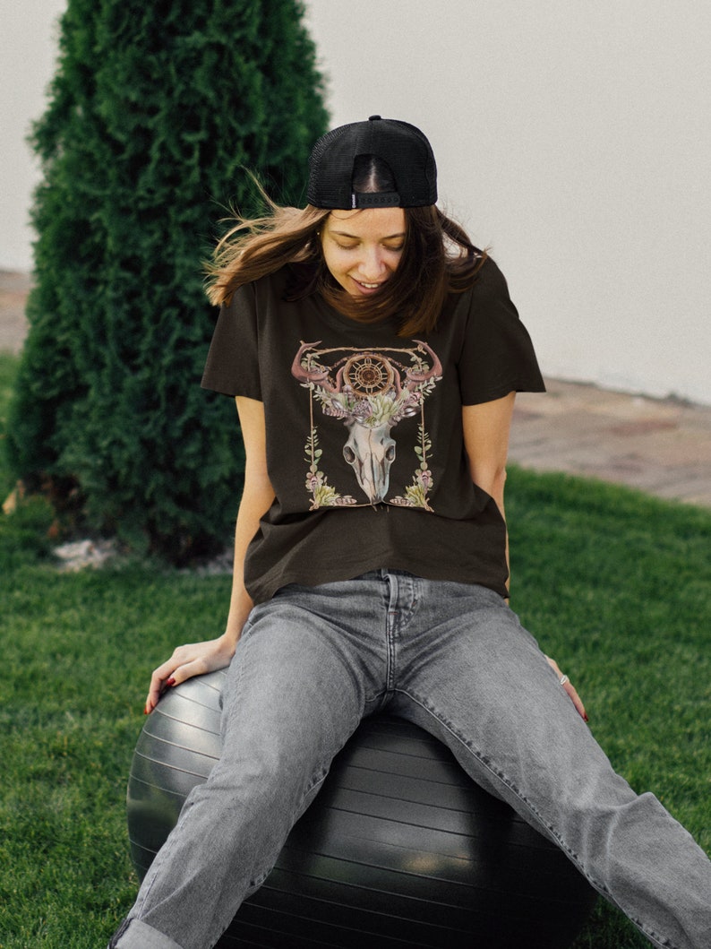 May include: A black t-shirt featuring a detailed graphic of a steer skull adorned with floral elements and a dreamcatcher design. The shirt is worn with gray jeans and a black baseball cap. The image is set outdoors with a green background.