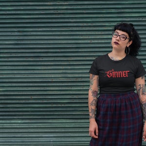 May include: A person wearing a black t-shirt with the word "Sinner" in red gothic lettering. The person has tattoos on their arms and is wearing a black and red plaid skirt.