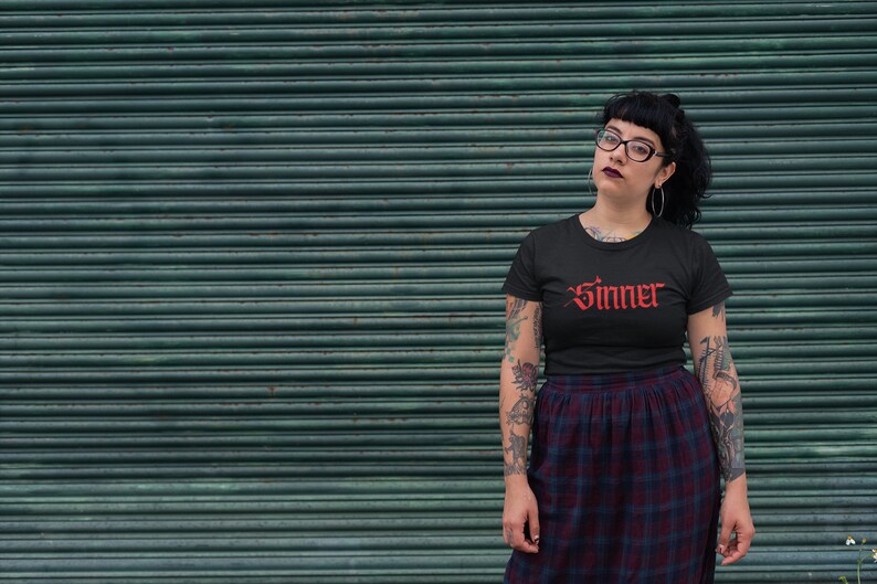 May include: A person wearing a black t-shirt with the word "Sinner" in red gothic lettering. The person has tattoos on their arms and is wearing a black and red plaid skirt.