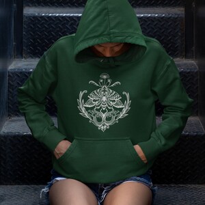 K&ouml;nnte beinhalten: Ein dunkelwaldgr&uuml;ner Kapuzenpullover mit einem wei&szlig;en Grafikdruck auf der Brust. Das Design zeigt einen zentralen Nachtfalter mit detaillierten Fl&uuml;gelmustern, flankiert von Blattzweigen und zwei schlangenartigen Figuren, gekr&ouml;nt von kleinen Pilzen.