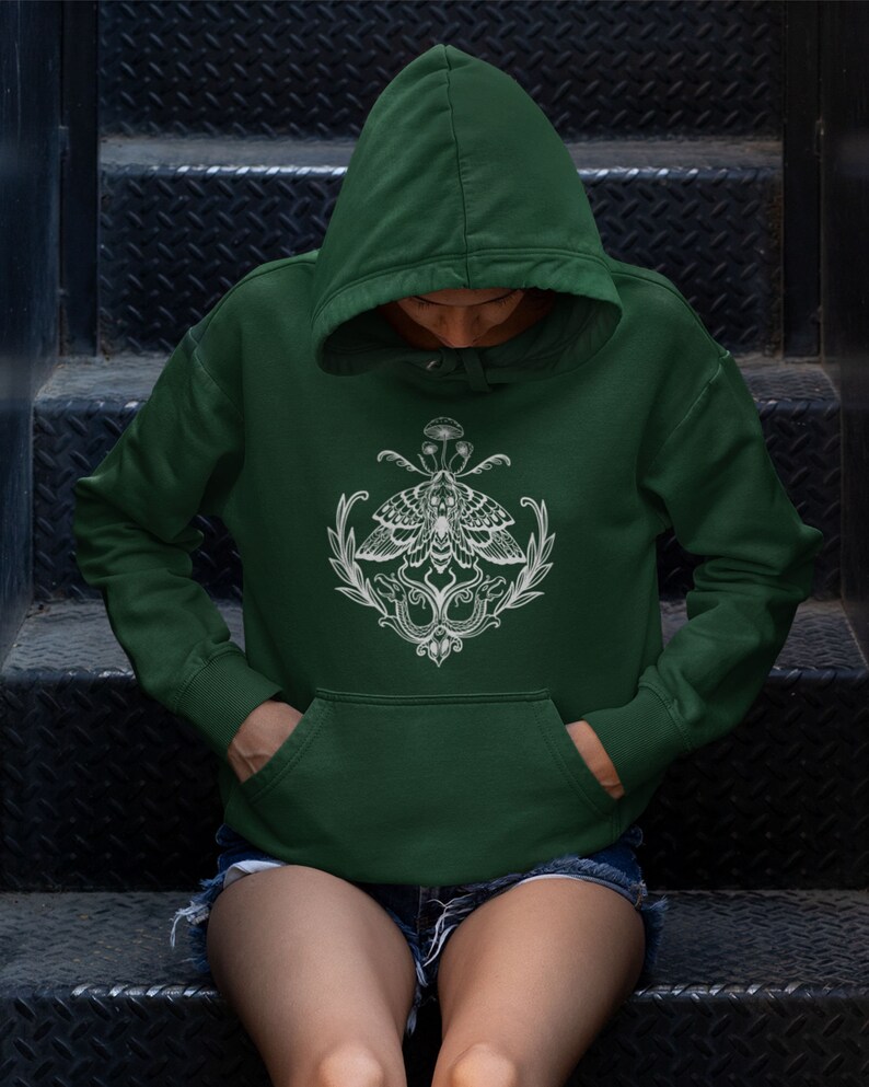 K&ouml;nnte beinhalten: Ein dunkelwaldgr&uuml;ner Kapuzenpullover mit einem wei&szlig;en Grafikdruck auf der Brust. Das Design zeigt einen zentralen Nachtfalter mit detaillierten Fl&uuml;gelmustern, flankiert von Blattzweigen und zwei schlangenartigen Figuren, gekr&ouml;nt von kleinen Pilzen.