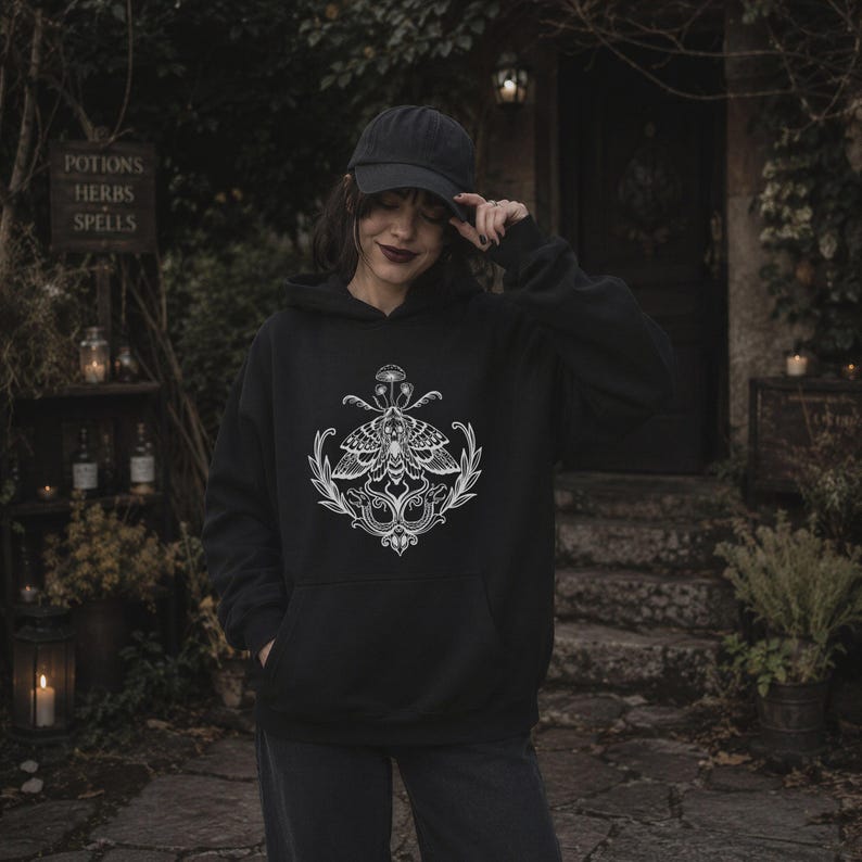 K&ouml;nnte beinhalten: Ein schwarzer Kapuzenpullover mit einem wei&szlig;en Grafikdruck auf der Brust. Das Design zeigt einen Nachtfalter &uuml;ber zwei Schlangen, die mit belaubten Zweigen verschlungen sind, gekr&ouml;nt von einem kleinen Pilz. Der Hoodie hat lange &Auml;rmel und eine Tasche.