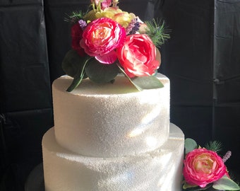 May include: Two-tiered white cake dummy with a floral topper. The topper features pink and yellow flowers with green leaves. The cake is on a silver cake stand.
