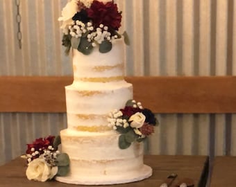 May include: A three-tiered white wedding cake with a rustic, naked cake design. The cake is decorated with burgundy and white flowers, greenery, and a gold topper.