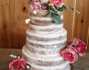 May include: A three-tiered naked wedding cake decorated with pink flowers and a wooden topper that says "E & F" with an arrow through a heart. The cake is on a silver cake stand and the background is a wooden wall.