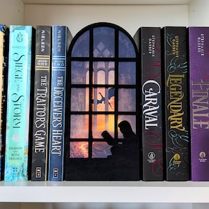 Peut inclure: Une étagère avec des livres variés, dont des titres comme "Shadow and Bone" et "Caraval." Un serre-livres décoratif en forme de fenêtre présente une silhouette d'une personne lisant. Les livres sont disposés sur une étagère blanche.