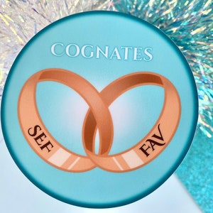 May include: A light blue round sticker with the words "Cognates" in white text at the top. Two gold rings are intertwined in the center of the sticker. The word "SEF" is inscribed on one ring and "FAV" is inscribed on the other ring.