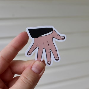 Könnte beinhalten: Ein gestanzter Aufkleber mit einer grafischen Illustration einer Hand mit fünf ausgestreckten Fingern, dargestellt in einem hellen Hautton mit schwarzen Umrissen. Die Hand ragt aus einem einfarbigen schwarzen Ärmelbündchen hervor.