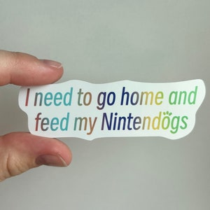 May include: A white sticker with a rainbow gradient text that reads "I need to go home and feed my Nintendogs".