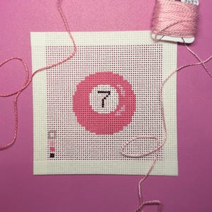 May include: A square needlepoint canvas featuring a pink billiard ball design with the number seven in the center. The canvas includes a small color key in the bottom left corner and is accompanied by a spool of pink embroidery thread.
