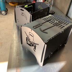 May include: Two black metal fire pits with cut-out designs. The fire pit on the bottom has a design of a soldier with a rifle and the text "Guard & Defend". The fire pit on top has a design of a soldier with a sword and the text "Guard & Defend".