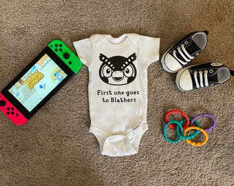 May include: A white baby onesie with a black owl graphic and the text "First one goes to Blathers". The onesie is next to a Nintendo Switch console with a green and red controller and a screen showing the Animal Crossing game. There are also two pairs of black and white baby shoes and a set of colorful teething rings.