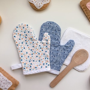 May include: Two blue and white patterned oven mitts, one with a floral design and the other with a geometric pattern, are displayed on a white surface with a wooden spoon, a white cotton potholder, and several small pastries.