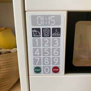May include: White toy microwave with a black door and a digital display showing 01:15. The control panel has buttons for numbers 1-9 and 0, as well as buttons for start, stop, and icons for popcorn, chicken, and a dish.