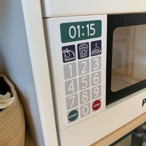 May include: White toy microwave with a black door and a digital display showing 01:15. The microwave has a keypad with numbers 1-9 and 0, as well as buttons for "Start" and "Stop". There are three icons above the keypad: a turkey, a popcorn bag, and a plate with a steaming dish.