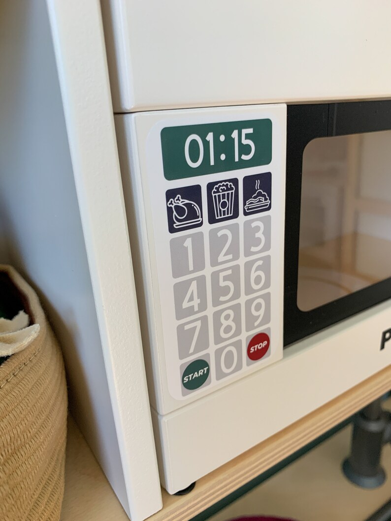 May include: White toy microwave with a black door and a digital display showing 01:15. The microwave has a keypad with numbers 1-9 and 0, as well as buttons for "Start" and "Stop". There are three icons above the keypad: a turkey, a popcorn bag, and a plate with a steaming dish.