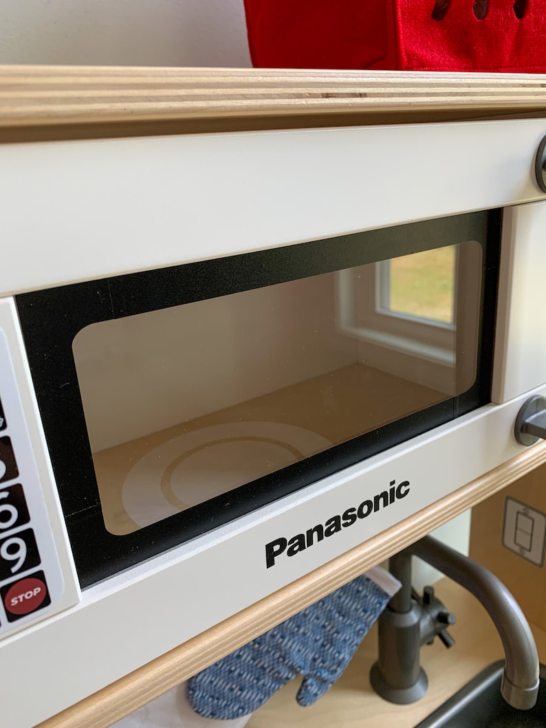 May include: White toy microwave with a black frame and a clear glass door. The microwave has a black Panasonic logo and a digital display with buttons labeled "Stop" and numbers 0-9.