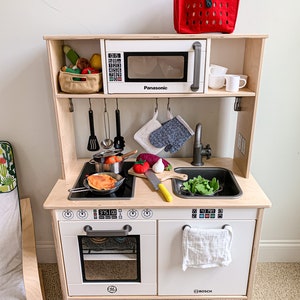 May include: A white wooden toy kitchen playset with a pretend oven, stovetop, sink, and microwave. The microwave has a Panasonic logo and a digital clock that reads 01:15. The oven has a Bosch logo and a digital display that reads 375. The sink has a faucet and a white towel hanging on the side. There are various toy kitchen accessories, including a pot, pan, utensils, and vegetables.