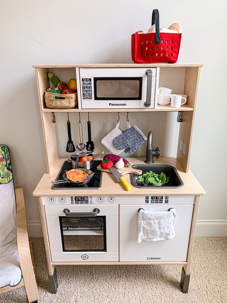 May include: A white wooden toy kitchen playset with a pretend oven, stovetop, sink, and microwave. The microwave has a Panasonic logo and a digital clock that reads 01:15. The oven has a Bosch logo and a digital display that reads 375. The sink has a faucet and a white towel hanging on the side. There are various toy kitchen accessories, including a pot, pan, utensils, and vegetables.