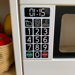 May include: White toy microwave with a black digital display showing 01:15. The microwave has a black keypad with numbers 1-9 and 0, as well as buttons for start and stop. There are three icons above the keypad: a chicken, popcorn, and a pie.