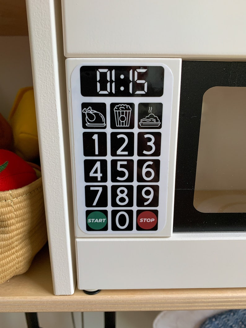 May include: White toy microwave with a black digital display showing 01:15. The microwave has a black keypad with numbers 1-9 and 0, as well as buttons for start and stop. There are three icons above the keypad: a chicken, popcorn, and a pie.