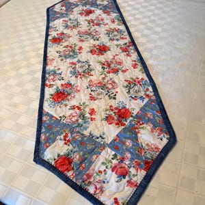 May include: A quilted table runner featuring a white background with a floral pattern of red and pink roses, green leaves, and blue accents. The runner has a pointed end and is bordered by a blue fabric with a white polka dot pattern.