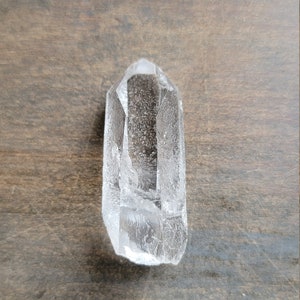 May include: A clear quartz crystal point with a natural, faceted shape. The crystal is resting on a brown wooden surface.